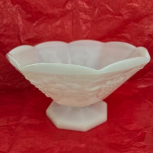 May include: A translucent white milk glass bowl featuring a scalloped rim and an embossed fruit pattern on the exterior. The bowl is supported by a hexagonal pedestal base, creating a footed design with a smooth, glossy finish throughout.
