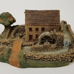 Puede incluir: Una casa de piedra en miniatura con una rueda de agua, un pequeño puente y un árbol. La casa es marrón y tiene un techo de piedra. La rueda de agua es de madera y está girando. El puente es de piedra y está sobre un pequeño arroyo. El árbol es verde y tiene ramas marrones.