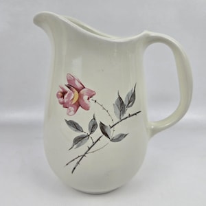 May include: Cream-coloured ceramic jug with a curved handle and spout. A pink rose with yellow accents and grey leaves adorns the front. The jug is a classic design, suitable for serving beverages.