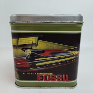 May include: A rectangular green metal tin with a silver lid. The tin features a black background with a red and yellow futuristic vehicle design. The text "A FUTURE DESIGN" and "FOSSIL" are printed on the front.