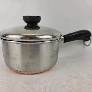 May include: A small stainless steel saucepan with a lid and a black handle. The pot has a copper-coloured base and a black knob on the lid. The handle has a metal ring at the end.