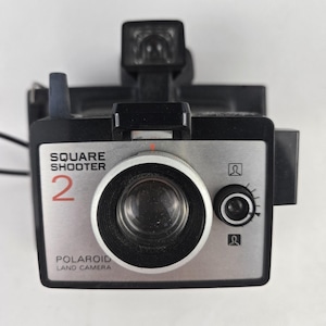 Puede incluir: Una cámara Polaroid Square Shooter 2 Land Camera vintage. La cámara es negra y plateada con una lente grande y un accesorio de flash. El texto "SQUARE SHOOTER 2" y "POLAROID LAND CAMERA" es visible en la parte delantera.