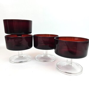 May include: Four vintage burgundy-colored glass dessert bowls. Each bowl has a wide, shallow, rounded top and a clear, stemmed base. The bowls are arranged on a white surface, showcasing their rich color and design.