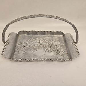 May include: A silver-colored metal serving tray with a handle. The tray features a floral rose design and a scalloped edge. The handle has a textured, branch-like appearance.