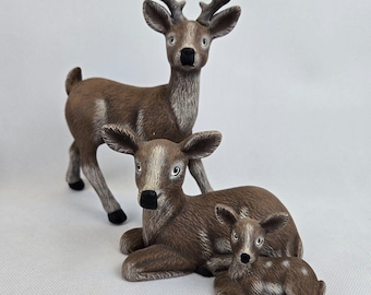 May include: A family of three ceramic deer figurines. The largest deer is standing with its head held high, while the two smaller deer are lying down. All three deer are brown with white markings.