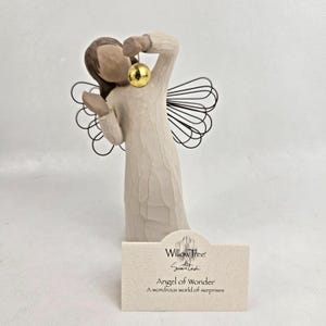 Puede incluir: Una figura de ángel color crema con alas de alambre sostiene un adorno dorado. La figura tiene una textura tallada similar a la madera y una paleta de colores neutros. Una pequeña tarjeta al frente dice "Angel of Wonder" y "A wondrous world of surprises."