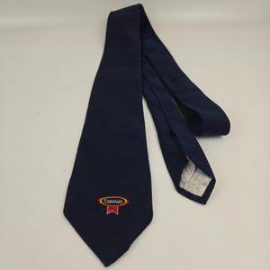 Può includere: Una cravatta blu navy con un logo ricamato "Colonial" in bianco e un design a nastro rosso. La cravatta è realizzata in tessuto strutturato. Viene presentata su una superficie bianca.
