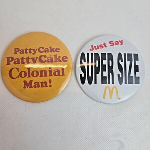 Può includere: Due bottoni rotondi vintage. Uno è giallo con il testo "Patty Cake Patty Cake Colonial Man!" in marrone. L'altro è bianco con "Just Say SUPER SIZE" in rosso e nero, con un logo "M" giallo.