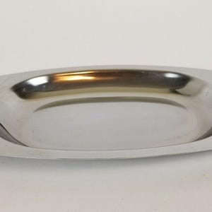 Puede incluir: Bandeja ovalada de acero inoxidable con asas doradas. La bandeja tiene una superficie pulida y reflectante y un diseño poco profundo y curvado. Las asas son rectangulares con un diseño similar al bambú.