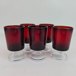 Può includere: Set di sei bicchieri a stelo rosso rubino. I bicchieri hanno un gambo corto e trasparente e una base arrotondata. La coppa di ogni bicchiere è di colore rosso intenso. I bicchieri sono disposti in gruppo su una superficie bianca.