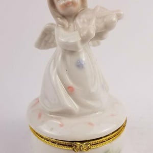 May include: A white porcelain trinket box with a hinged lid. The box features a figurine of an angel playing a violin. The angel has a white dress with pink and blue accents. The box is decorated with yellow flowers and green leaves.