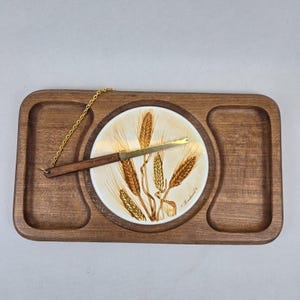 Op de afbeelding: Een houten kaasplank met een rond keramiek bord met een tarweontwerp. Het bord heeft een klein mes dat eraan vastzit met een ketting.