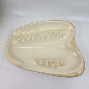 May include: A white ceramic ashtray with two indented areas for cigarettes. The ashtray is shaped like a leaf and has a slightly textured surface.