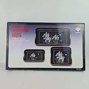 Puede incluir: Un juego de tres bandejas lacadas en negro con ribetes dorados, con diseños florales de iris blancos. La caja dice "WHITE IRIS FINEST 3PCS LACQUERED TRAY". Las bandejas son de diferentes tamaños, con asas doradas.