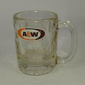 May include: A clear glass A&W mug with a handle. The mug features the A&W logo in orange and brown. The mug has a textured design on the exterior and is empty.