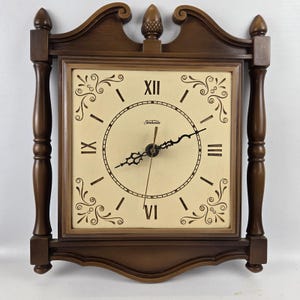 May include: A vintage brown wooden wall clock with a square face and decorative scrollwork. The clock face is cream-colored with black Roman numerals and hands. The clock has a decorative top and side supports.