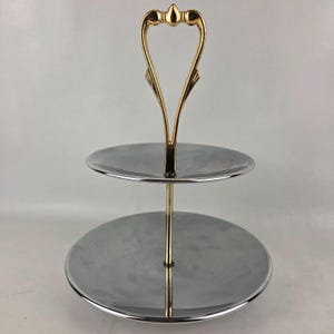 May include: Two-tiered serving tray with a polished silver finish and a gold-colored handle. The handle has a decorative, heart-shaped design. The tray is suitable for serving desserts, appetizers, or other small items.