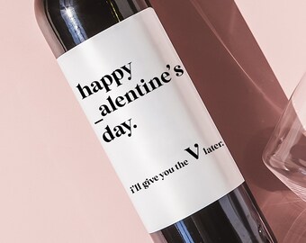 May include: A bottle of red wine with a white label that says "happy valentine's day. I'll give you the V later." The bottle is on a pink surface with two wine glasses.