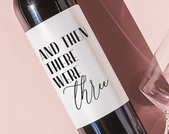 May include: A bottle of red wine with a white label that says "And then there were three". The bottle is on a pink surface with two wine glasses.