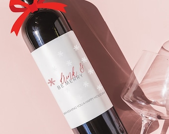 May include: A bottle of red wine with a white label that says "Drink & Be Merry" and a red bow. The bottle is on a pink surface with two wine glasses.
