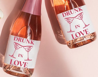 May include: Three mini bottles of pink sparkling wine with labels that say "Drunk in Love" and a pink floral arrangement on a pink background.