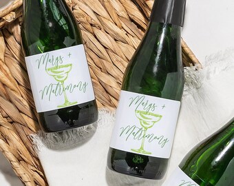 May include: Three small glass bottles with black caps and white labels. The labels have green text that says "Mimosas +" and a green illustration of a margarita glass. The bottles are in a woven basket with a white cloth.