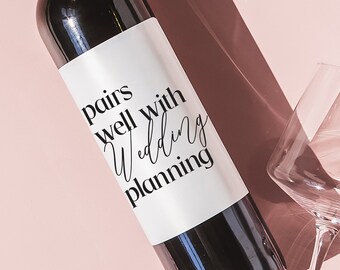 May include: A dark wine bottle with a white label that reads "pairs well with Wedding planning" is positioned on a pink surface. Two wine glasses are also present, casting shadows. The image has a soft, natural lighting.