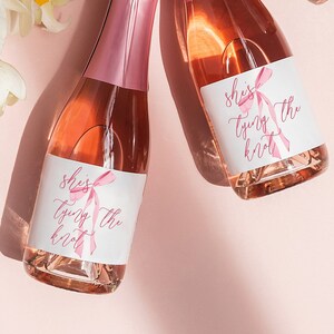 May include: Three mini bottles of pink sparkling wine with white labels that say "She's tying the knot" in pink script with a pink ribbon graphic. The bottles are on a pink background with white orchid flowers.