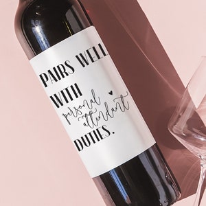 May include: A bottle of red wine with a white label that says "Pairs well with personal attendant duties." The bottle is on a pink surface with two wine glasses.