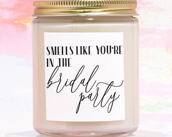 May include: A clear glass candle jar with a gold lid and a white label that reads "Smells like you're in the bridal party". The candle is sitting on a clear acrylic block.