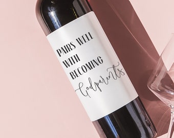 May include: A dark red wine bottle with a white label that reads "Pairs Well With Becoming Godparents" is displayed on a pink background. Two clear wine glasses are positioned around the bottle, with shadows cast across the surface.