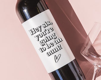May include: A bottle of red wine with a white label that says "Hey sis, you're going to be an aunt!" with a small heart symbol. The bottle is on a pink surface with two wine glasses.