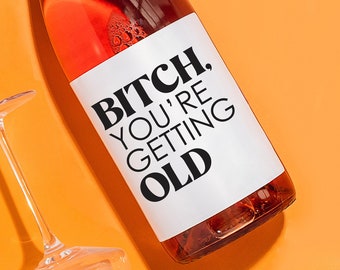 May include: A bottle of rosé wine with a silver cap and a white label that reads "BITCH, YOU'RE GETTING OLD" in black text. The bottle is surrounded by four clear wine glasses on an orange background.