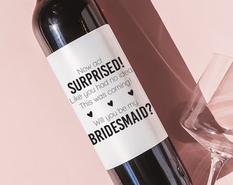 May include: A white wine bottle with a black label that says "Now act SURPRISED! Like you had no idea This was coming! Will you be my BRIDESMAID?"
