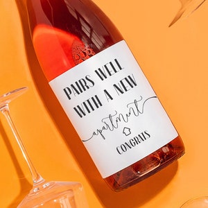 May include: A bottle of rose wine with a white label that says "Pairs well with a new apartment" and "Congrats". The bottle is surrounded by four clear wine glasses on a bright orange background.