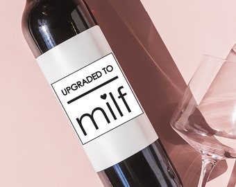 May include: A bottle of red wine with a white label that says "Upgraded to MILF" with a small heart symbol. The bottle is on a pink surface with two wine glasses.