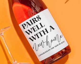 May include: A bottle of rose wine with a white label that says "Pairs well with a new home. Congrats on the new place!" The bottle is surrounded by four clear wine glasses on a bright orange background.