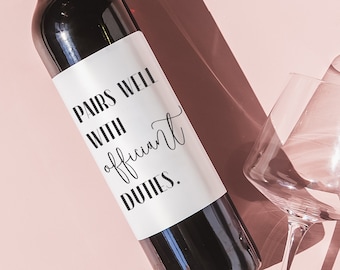 May include: A dark red wine bottle with a white label that reads "Pairs well with officiant duties." Two wine glasses are positioned around the bottle on a light pink background. The image has a shadow.