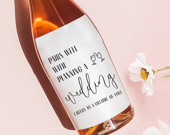 May include: A pink wine bottle with a white label that says "Pairs well with planning a wedding. Cheers to a lifetime of love." The bottle is surrounded by white flowers.
