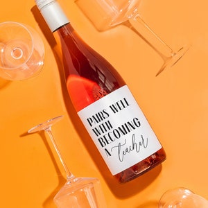 May include: A bottle of rose wine with a white label that says "Pairs well with becoming a teacher". The bottle is surrounded by four empty wine glasses on a bright orange background.
