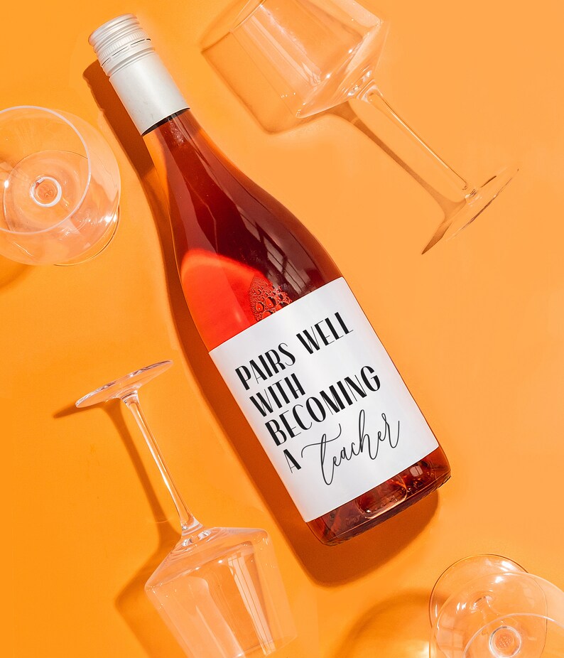 May include: A bottle of rose wine with a white label that says "Pairs well with becoming a teacher". The bottle is surrounded by four empty wine glasses on a bright orange background.