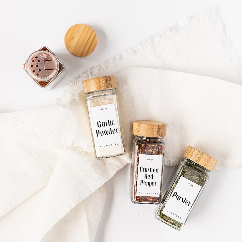 May include: Three glass spice jars with bamboo lids and white labels. The labels have black text that reads "Garlic Powder", "Crushed Red Pepper", and "Parsley". All labels have the text "ALL NATURAL" and a number.