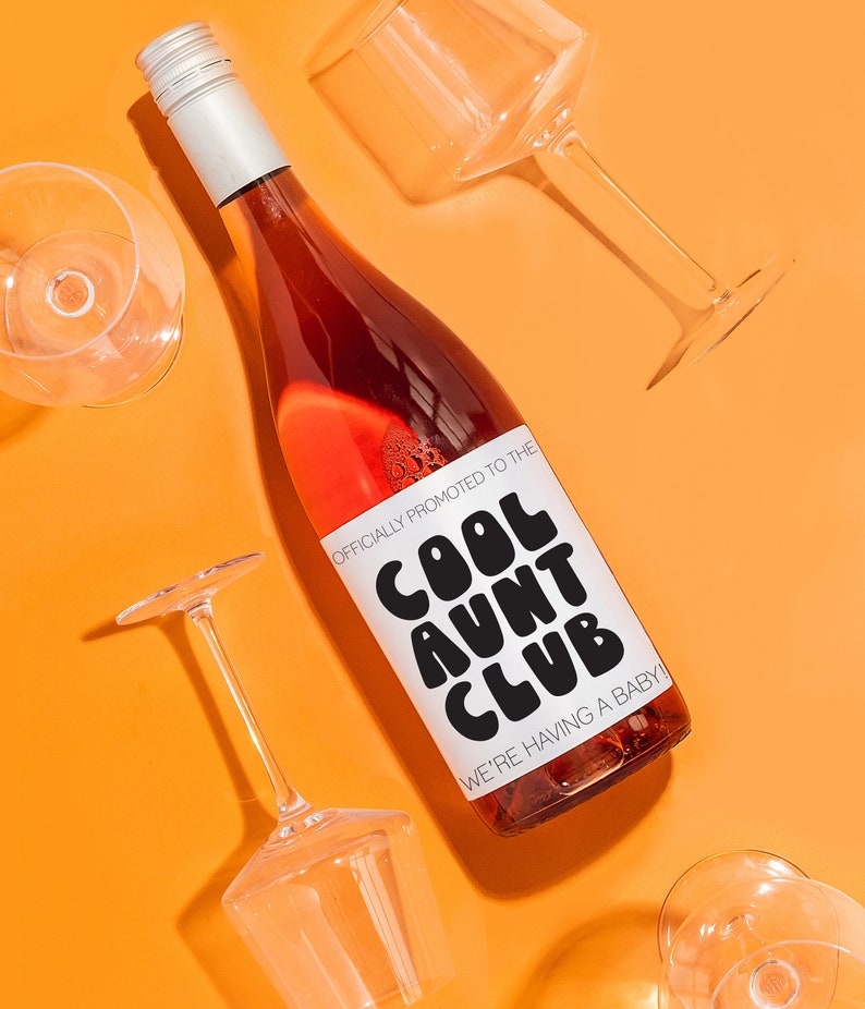May include: A bottle of rose wine with a white label that says "Officially Promoted to the Cool Aunt Club We're Having a Baby!" The bottle is surrounded by four clear wine glasses on a bright orange background.