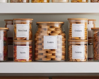 May include: A white shelf with six glass jars with bamboo lids. The jars are filled with various snacks, including cookies, crackers, cashews, dried fruit, and sweets. Each jar has a white label with black text indicating the contents. The labels read "Cookies", "Crackers", "Cashews", "Dried Fruit", and "Sweets".