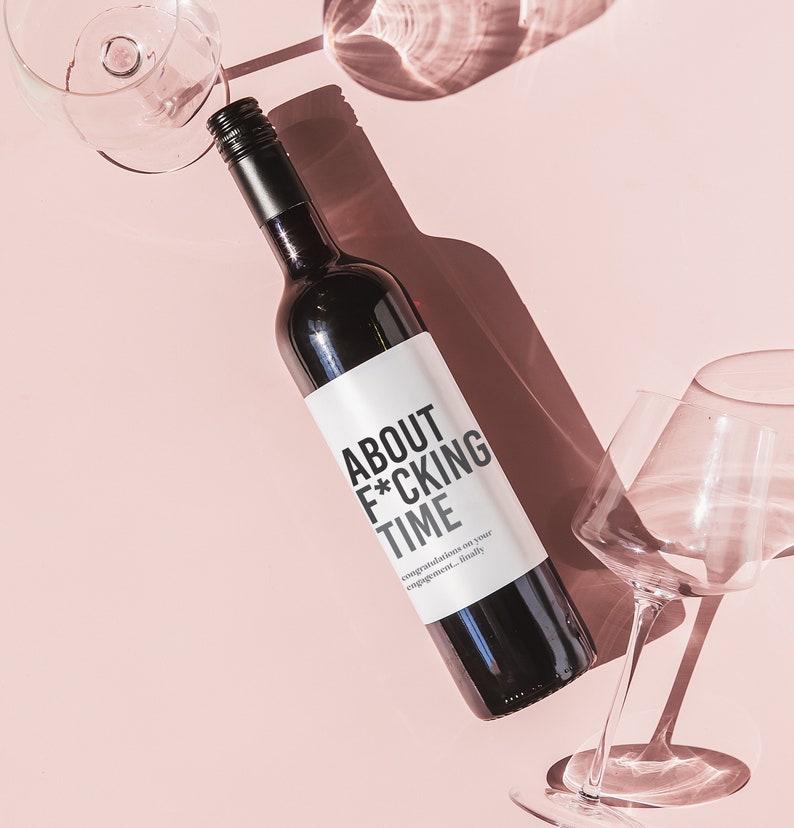 Może przedstawiać: Butelka czerwonego wina z białą etykietą, na kt&oacute;rej widnieje napis "About F*cking Time" i "Congratulations on your engagement...finally". Butelka stoi na r&oacute;żowym tle z dwoma kieliszkami do wina.