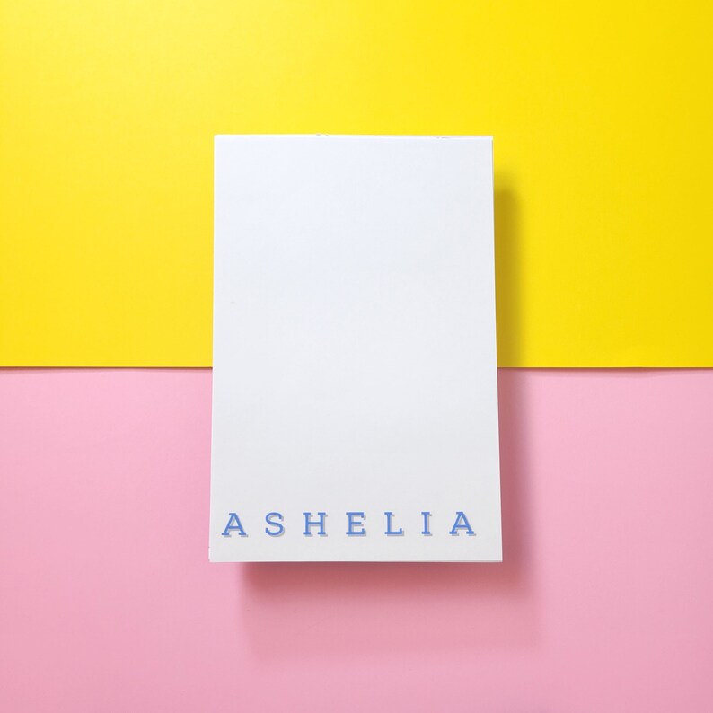 May include: A white card with the name "ASHELIA" printed in blue letters on a yellow and pink background.