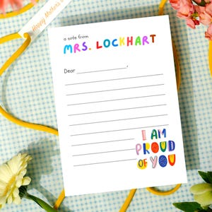 Può includere: Un foglio di carta da lettere bianco a righe con la scritta "a note from MRS. LOCKHART" in lettere colorate. La frase "I AM PROUD OF YOU" è stampata in basso. Un nastro giallo e fiori sono sullo sfondo.