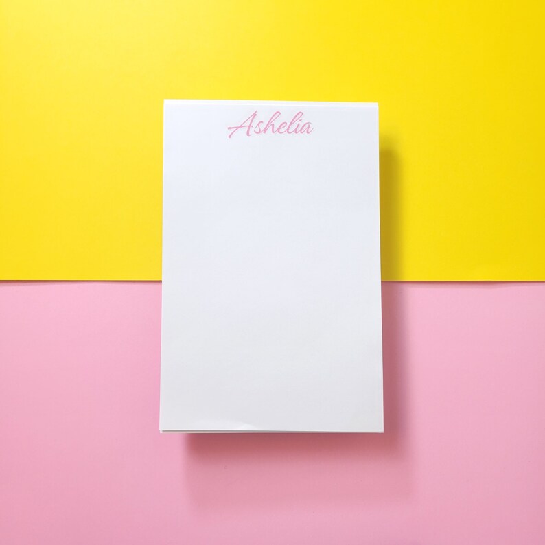 Op de afbeelding: Een wit notitieboekje met de naam "Ashelia" in roze bovenaan. Het notitieboekje ligt op een gele en roze achtergrond.