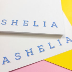 May include: Two white notepads with the name "SHELIA" printed in blue on the top notepad and "ASHELIA" printed in blue on the bottom notepad.