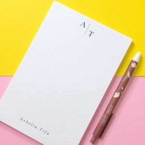 Puede incluir: Un bloc de notas blanco con las iniciales "A|T" y el nombre "Ashelia Tifa" impreso en la parte inferior. Un bolígrafo marrón con diseños florales está sobre una superficie rosa junto al bloc de notas.